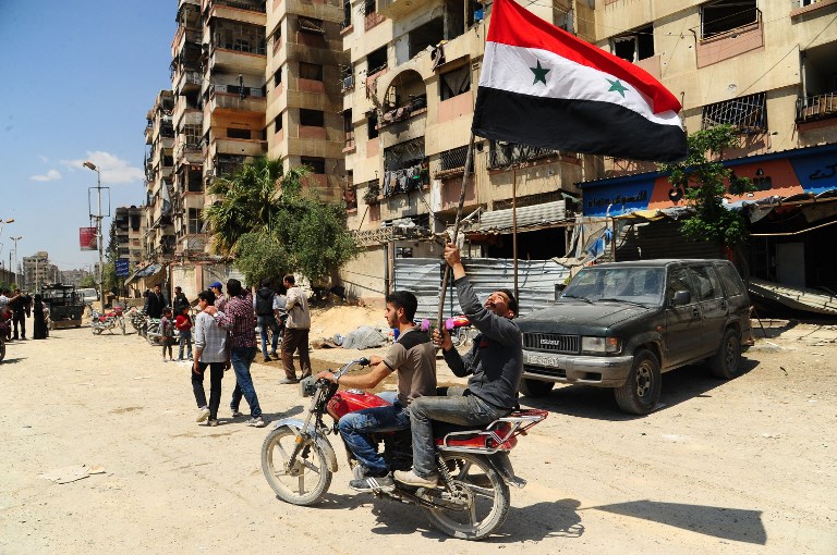 A handout picture released by the official Syrian Arab News Agency (SANA) on April 14, 2018, shows men waiving the Syrian flag as they drive a motorcycle in a street in the Eastern Ghouta town of Douma after Syrian government forces entered the last rebel bastion.