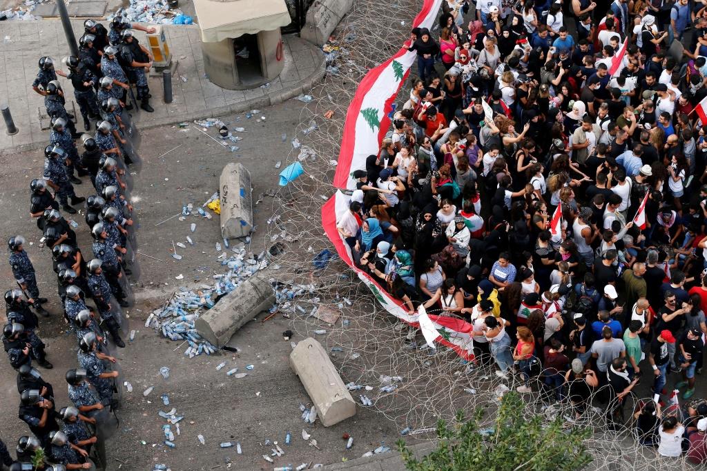 Demonstrators carry national flags as riot police stand guard behind barbed wire during an anti-government protest in Beirut, Lebanon October 19, 2019.